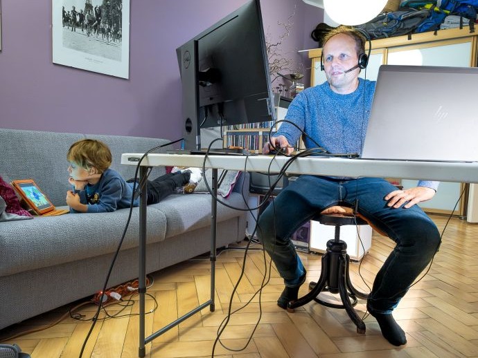 Ein Mann sitzt zu Hause am Computer; ein Kind liegt auf dem Sofa daneben, ebenfalls vor einem Bildschirm. Ein Mann sitzt zu Hause am Computer; ein Kind liegt auf dem Sofa daneben, ebenfalls vor einem Bildschirm.